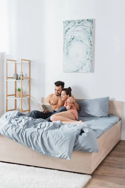 Alegre pareja sentado en gris ropa de cama y lectura libro - foto de stock