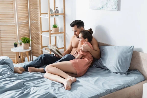 Joven hombre sin camisa abrazando novia en camiseta mientras lee libro en dormitorio - foto de stock