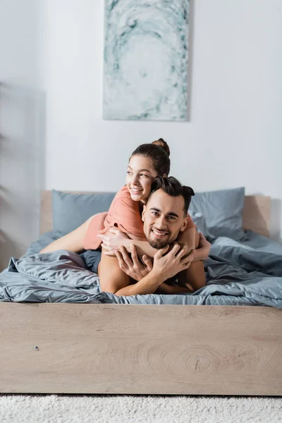 Happy man looking at camera near girlfriend hugging him in bedroom — Stock Photo