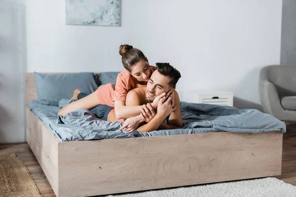 Femme séduisante étreignant jeune homme souriant couché sur la literie grise — Photo de stock