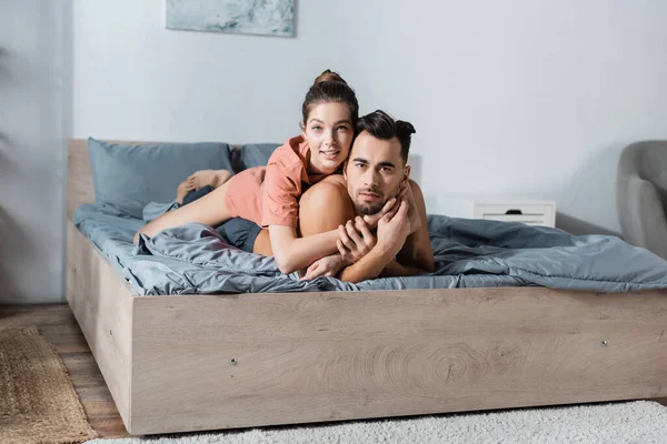 Junges und sexy Paar lächelt in die Kamera und umarmt sich auf grauer Bettwäsche — Stockfoto