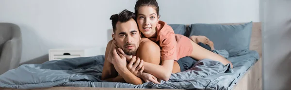 Junges Liebespaar, das sich auf dem Bett umarmt und in die Kamera blickt, Banner — Stockfoto