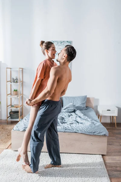 Side view of muscular shirtless man raising up amazed girlfriend in bedroom — Stock Photo