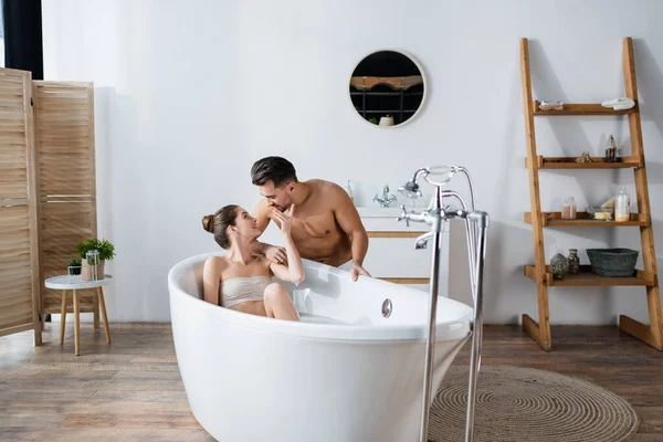 Sensual woman relaxing in bathtub while touching face of young shirtless boyfriend — Stock Photo