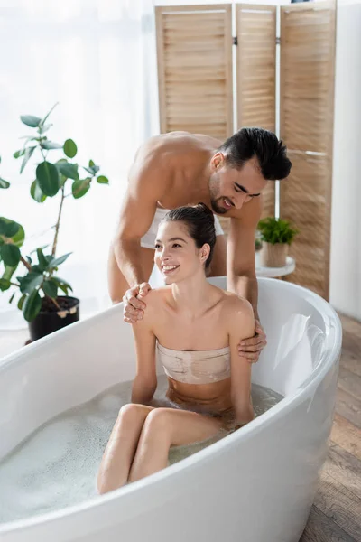 Lächelnder, hemdsloser Mann, der Schultern einer glücklichen Freundin berührt, die zu Hause baden geht — Stockfoto