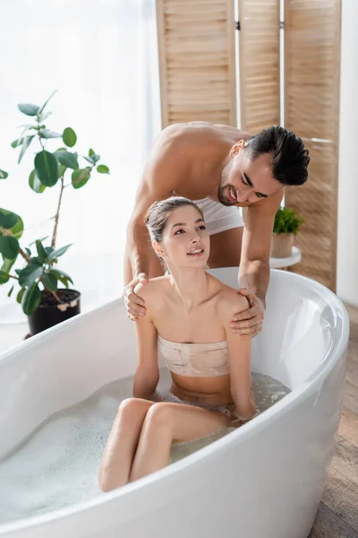 Улыбающийся мужчина без рубашки обнимает плечи сексуальной женщины принимающей ванну дома — стоковое фото