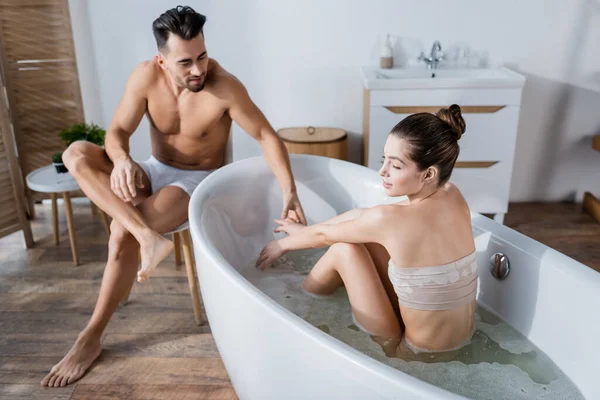 Young seductive woman taking bath and holding hands with shirtless man in underpants — Stock Photo