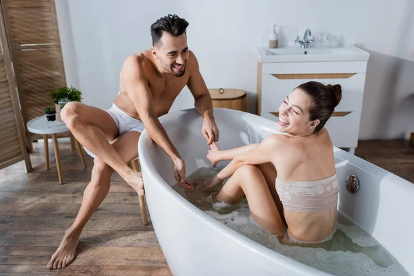 Mujer emocionada riendo en la bañera cerca de novio sonriente sentado en calzoncillos - foto de stock