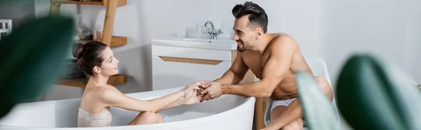 Young woman in bathtub and sexy shirtless man holding hands and looking at each other, banner — стоковое фото