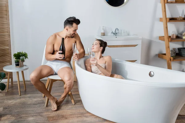 Jeune femme en baignoire et homme sexy en caleçon tenant des verres de champagne et se souriant — Photo de stock