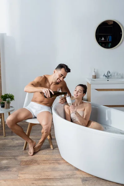 Hombre feliz en calzoncillos vertiendo champán en una copa de mujer relajándose en la bañera - foto de stock