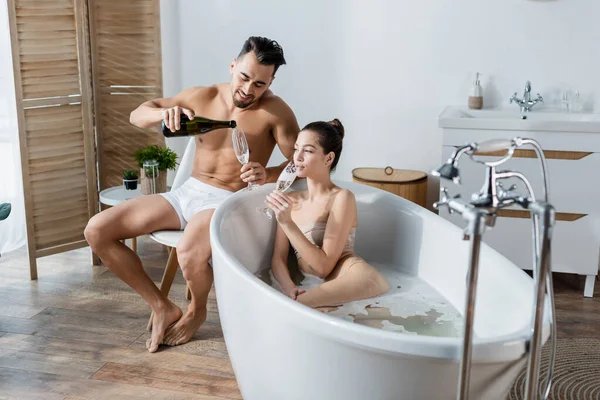 Homme souriant en caleçon versant champagne près de petite amie prendre un bain dans la salle de bain spacieuse — Photo de stock