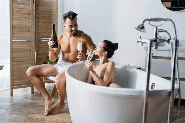 Feliz y sexy pareja sosteniendo copas de champán y hablando en el baño - foto de stock
