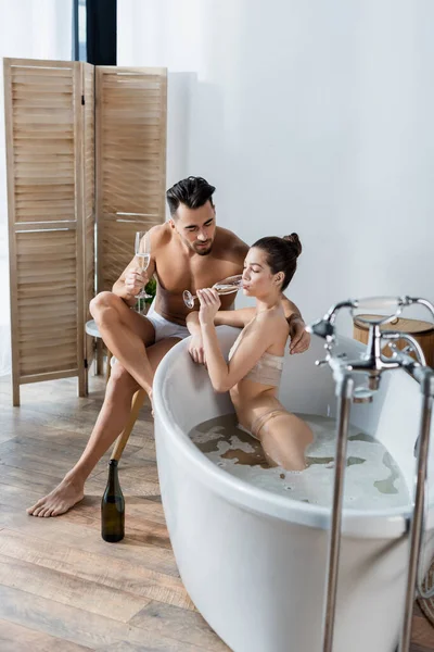 Verführerische Frau trinkt Champagner in Badewanne neben Freund in Unterhose — Stockfoto