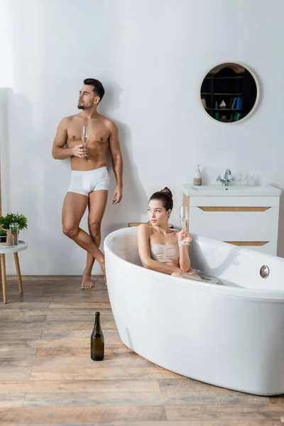 Sexy hombre en calzoncillos de pie con copa de champán cerca de novia relajarse en la bañera - foto de stock