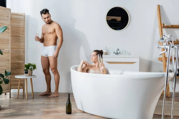 Femme souriante se détendre dans la baignoire près du petit ami en caleçon debout avec verre de champagne — Photo de stock