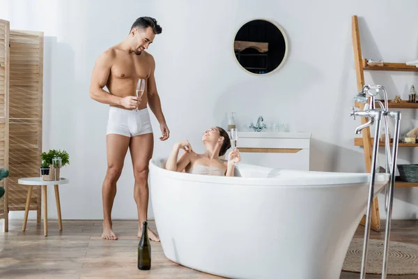 Mujer feliz relajándose en la bañera y mirando al novio en calzoncillos de pie con copa de champán - foto de stock
