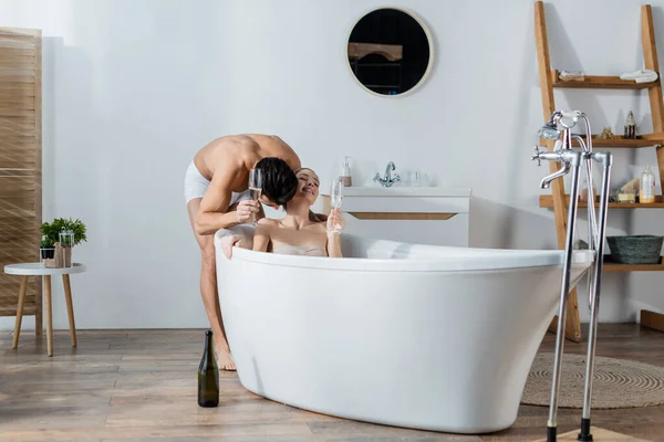 Jovem segurando taça de champanhe e beijando sorridente namorada relaxante na banheira — Fotografia de Stock