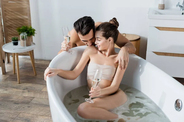 Shirtless man kissing shoulder of seductive girlfriend holding champagne glass in bathtub — Stock Photo