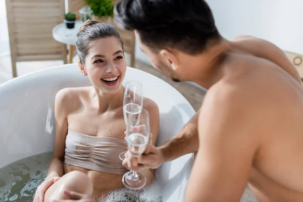 Fröhliche Frau badet und blickt ihren verschwommenen Freund in der Nähe von Sektgläsern an — Stockfoto