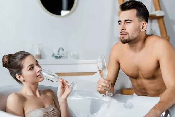 Sexy mujer bebiendo champán en bañera y mirando pensativo novio - foto de stock