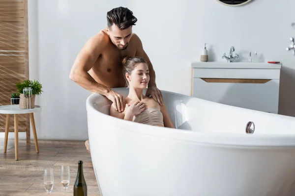 Sexy shirtless man massaging shoulders of girlfriend relaxing in bathtub with closed eyes — стоковое фото