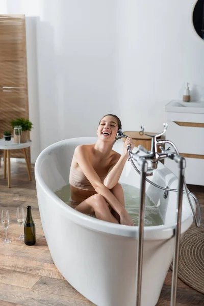 Смеющаяся женщина, принимающая ванну и имитирующая телефонный разговор с душевой головой возле бокалов и бутылки шампанского — стоковое фото