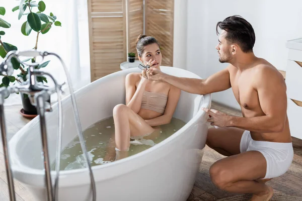 Muskulöser Mann in Unterhose zündet sich Zigarette von Freundin an, die sich in Badewanne entspannt — Stockfoto