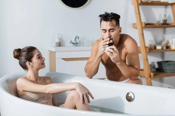 Femme séduisante souriante prenant un bain et regardant un homme torse nu allumer une cigarette — Photo de stock
