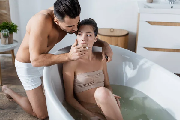 Sexy man in underpants holding cigarette near sensual woman taking bath at home — стоковое фото