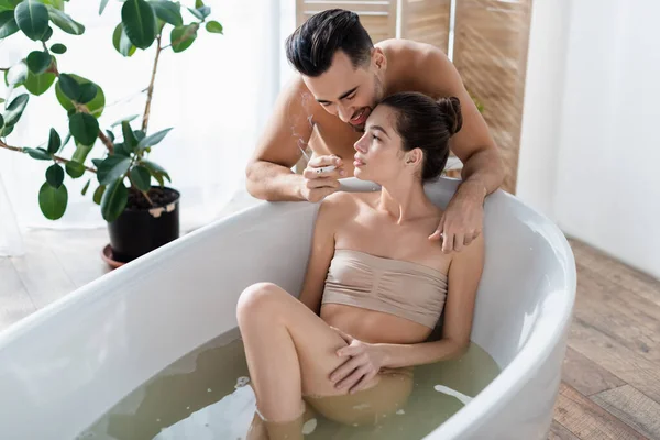 Smiling man holding cigarette near sexy girlfriend taking bath at home — Stock Photo