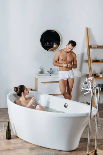 Sexy man in underpants lighting cigarette near girlfriend relaxing in bathtub with glass of champagne — Stock Photo