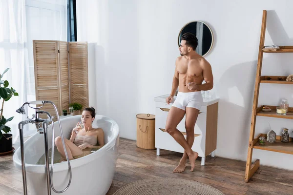 Young seductive woman drinking champagne in bathtub near sexy man holding cigarette in bathroom — Stock Photo