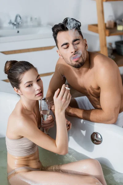 Joven sensual mujer en bañera celebración de cigarrillo y champán copa cerca sexy novio - foto de stock