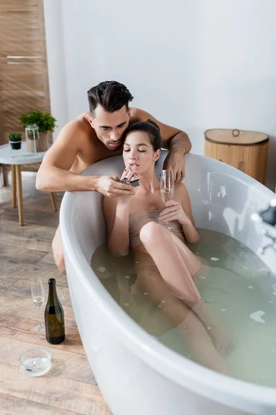 Young man lighting cigarette of girlfriend holding champagne glass while taking bath at home — Stock Photo