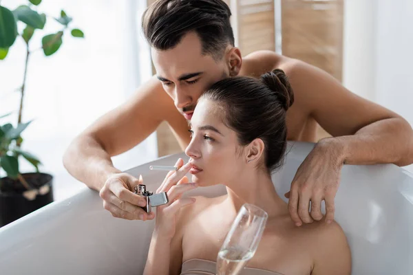 Shirtless man lighting cigarette of girlfriend taking bath and holding glass of champagne — Stock Photo