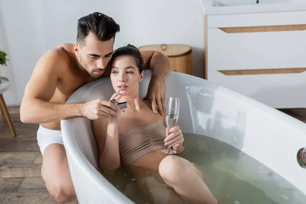 Sexy mujer sosteniendo champán mientras toma baño cerca de hombre encendiendo su cigarrillo - foto de stock