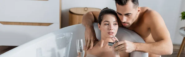 Shirtless man lighting cigarette of girlfriend relaxing in bathtub with champagne glass, banner — Stock Photo