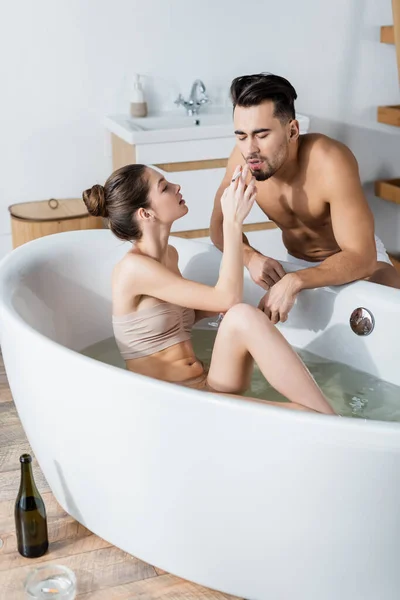 Sensual woman taking bath and holding cigarette near boyfriend with muscular torso — Stock Photo