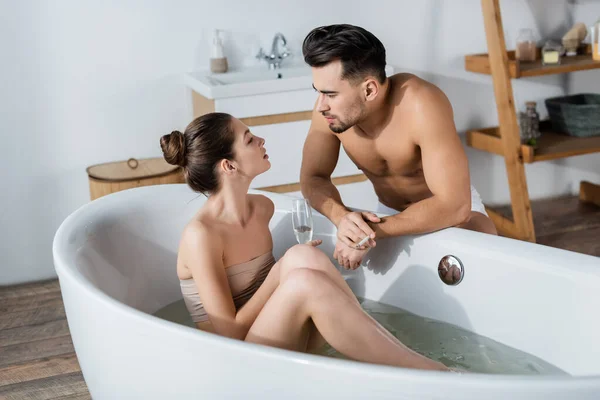 Сексуальная женщина с бокалом шампанского принимает ванну и смотрит на парня без рубашки в ванной — стоковое фото