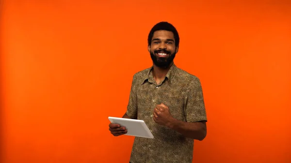 Joven afroamericano positivo con barba sosteniendo tableta digital sobre fondo naranja - foto de stock