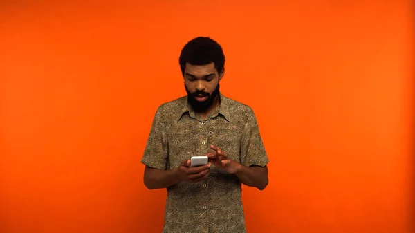 Choqué homme afro-américain messagerie sur smartphone sur fond orange — Photo de stock