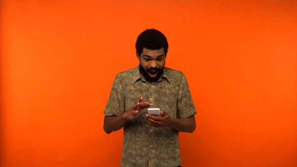 Espantado homem americano africano usando smartphone em fundo laranja — Fotografia de Stock