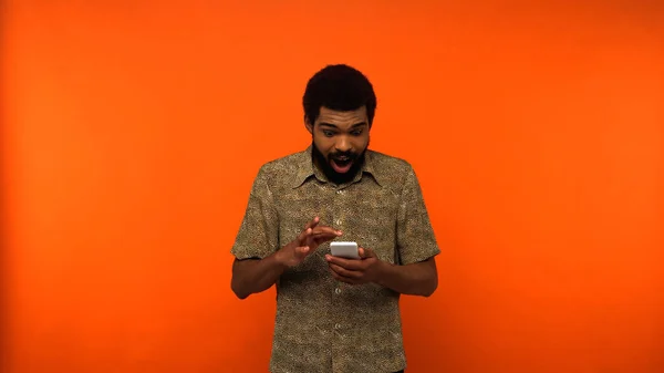 Hombre afroamericano asombrado usando teléfono inteligente sobre fondo naranja — Stock Photo