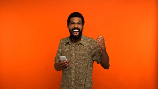 Hombre afroamericano emocionado con barba sosteniendo teléfono inteligente sobre fondo naranja - foto de stock