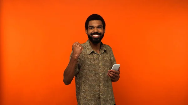 Hombre americano africano alegre con barba sosteniendo teléfono inteligente sobre fondo naranja - foto de stock