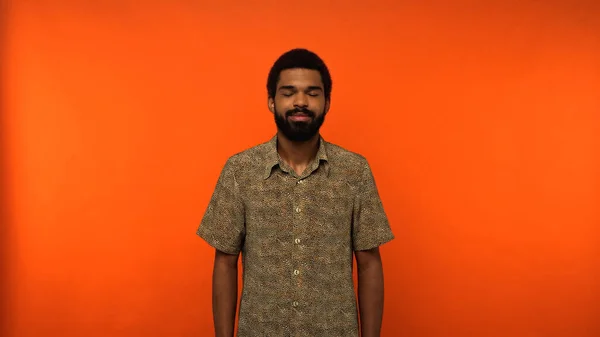 Homme afro-américain barbu et heureux avec les yeux fermés debout en chemise sur fond orange — Photo de stock