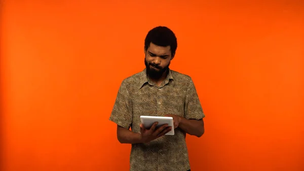 Спантеличений афроамериканський молодий чоловік з бородою тримає цифровий планшет на помаранчевому фоні — стокове фото