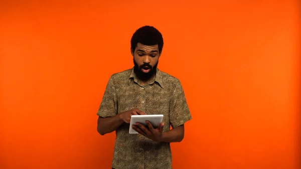 Удивленный африканский американский молодой человек с бородой, держащий цифровой планшет на оранжевом фоне — стоковое фото