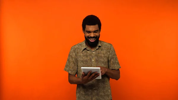 Fröhlicher afrikanisch-amerikanischer junger Mann mit Bart, der ein digitales Tablet auf orangefarbenem Hintergrund hält — Stockfoto